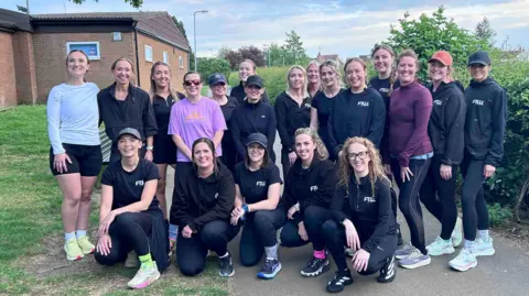 BBC/ Kate Bradbrook Nineteen runners, mostly wearing black tops and leggings, stand and kneel in two rows on a path, posing for the camera. 