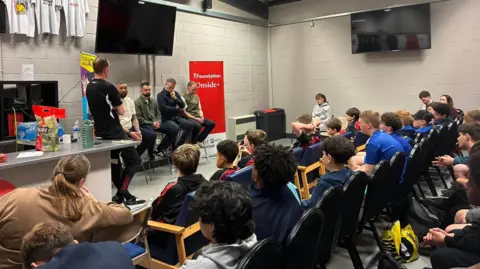 A group of children sit in black and blue chairs listening to a panel of five men including Tony Bellew and Jack McMullen