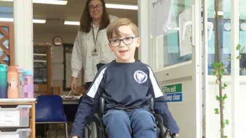 Jamie Niblock/BBC Ernie wearing his navy school uniform navigating his wheelchair through an open door. In the background his teacher wears a white shirt and is smiling at him from inside the classroom.