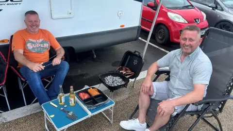 Alwyn Darragh, right, pictured with Ian Milligan who is on the left of the image. Both men are in deck chairs and sitting outside a camper van. They have a bbq on th go, which is sitting beside a fold up table on which there is food and drinks. Alwyn is smiling, and wearing a grey shirt and shorts. ian is wearing an orange t shirt and denim jeans