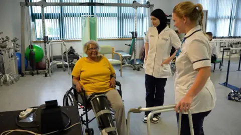A woman wearing a yellow t-shirt is sat in a wheelchair and smiles up at two nursing staff.