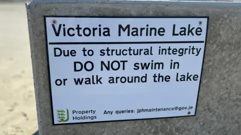 A white Government of Jersey warning signed screwed into a giant piece of stone at Victoria Marine Lake. The sign advises people not to swim in or walk around the lake. 