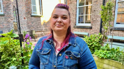 DAVE EDWARDS/BBC A woman looks at the camera. She has dyed pink hair and a neutral expression.