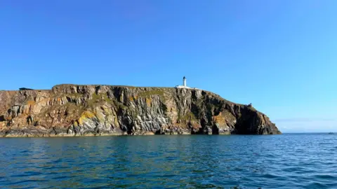 A view of the Mull of Galloway with a blue sky above, rocky coastline, a lighthouse and water all around