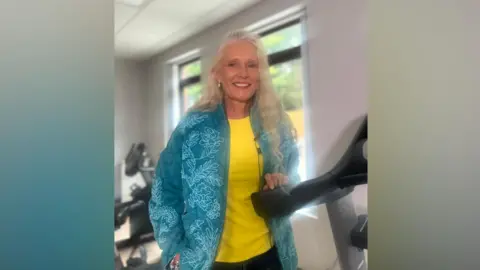 A woman smiling at the camera. She has long grey hair and is wearing a yellow top and blue jacket.