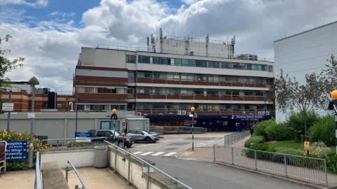 The outside of a hospital building. It is at least five storeys high, and there is a road leading up to it. There is a blue sky and clouds above.
