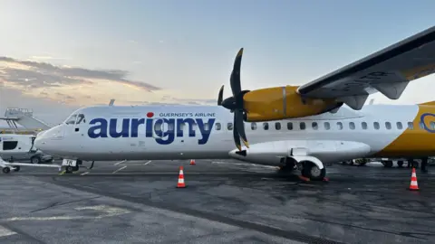 The picture shows an Aurigny aircraft parked on an airport apron. The plane has two large propellers, one of which is clearly visible near the wing. The fuselage is white with the word “aurigny” written in bold blue letters, and underneath it reads “Guernsey’s Airline.” The tail section and engine housing are painted in a bright yellow colour. There are orange traffic cones placed near the aircraft, and the ground appears to be a tarmac surface. In the background, there is airport equipment and a partly cloudy sky.