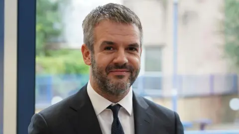 BBC/Oli Constable Coppard wears a black suit and tie as he smiles at the camera. He has salt and pepper hair and facial stubble.