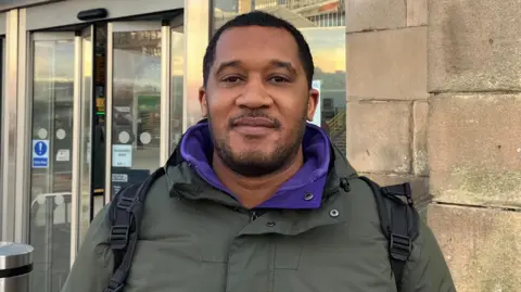 Simon Coombs stood outside Sheffield train station. He has short, dark hair and is wearing a green jacket with a purple sweater underneath.