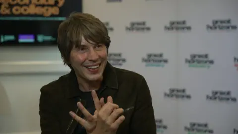 Professor Brian Cox sits in front of a white background. He wears a black jacket and a black T-shirt. He has collar-length dark hair and a sweeping fringe.