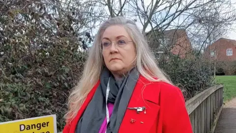 Darren Calpin/LDRS Julie Stevenson who has long grey/blonde hair and is wearing rimless glasses, a red coat over a grey scarf and pink top. She is standing on a wooden bridge with houses glimpsed behind her through trees/shrubs. 