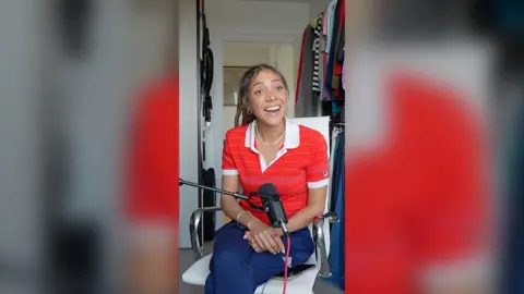 Miquita Oliver wears a red polo shirt and sits in a white office chair with a mic in front of her. She is smiling.