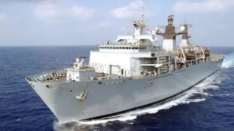 MOD HMS Bulwark is shown sailing on the open ocean, with its grey hull, superstructure, and various antennas and structures visible. The ship appears to be underway, creating a wake as it moves.