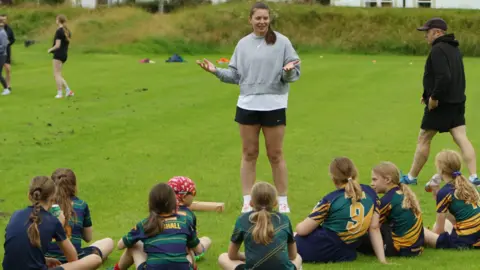 Young rugby players sitting on the grass dressed in green and yellow tops being inspired by England's Abbie Ward