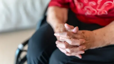 A stock image of an elderly lady grasping her hands together. She is sitting in a wheelchair.