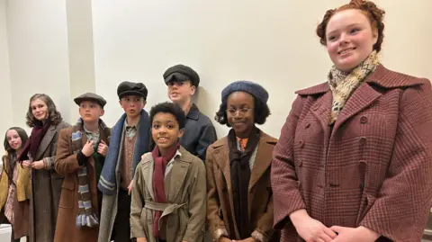Eight children in 1940s costumes standing in a hallway.