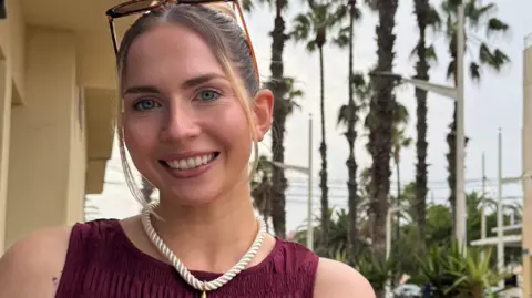 Submitted A woman smiles at the camera - she has her blonde hair tied back but two strands frame her face. She has blue eyes and sunglasses on top of her head. She is wearing a sleeveless maroon dress and a necklace which resembles white-coloured rope. Palm trees and a pale-coloured building are behind her.