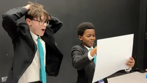 Two boys in school uniform, one pretending to paint a picture, the other protraying amazement at the results.