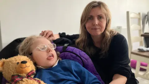 Mark Ansell/BBC Dee Cowburn pictured with her daughter Tilly. Tilly, who has blonde hair, is positioned next to a teddy bear. Dee, who also has blonde hair, stares directly at the camera with a neutral expression.