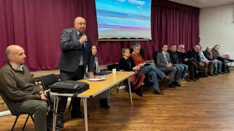 A group of council officials and councillors, and coastal management experts sit in a row before a stage at a meeting in Hemsby Village Hall. Carl Smith, the council leader is standing at a table, holding a microphone. A screen has a projected image on it, mounted on a backdrop of a burgundy coloured curtain.