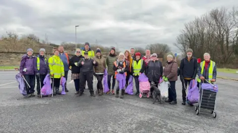 A group of eighteen people are wearing hi-vis vests and are carrying purple bin bags as part of the litter pick. They are standing in a car park which is surrounded by trees. 