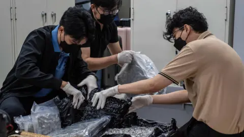 Jonathan Head/BBC Three men in masks search a suitcase full of vacuum packed black and see-through bags