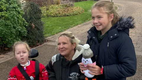 Two children either side of their mother. The younger child is in a wheelchair. The older child is standing up and holding a penguin soft toy.