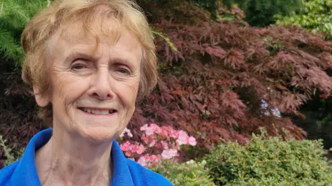 Anne Cobham with short light brown hair wearing a blue top stands in the Botanic Gardens on a sunny day with foliage and flowers behind her. She is smiling.