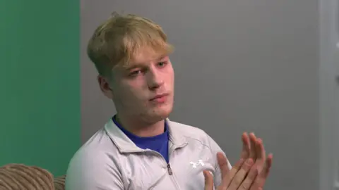 A blonde-haired teenage boy with a grey zip up jumper on, rubbing his hands together whilst looking into the distance.