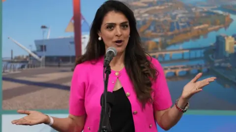 PA Media Natasha Asghar speaking into a microphone mounted on a stand. She is wearing a bright pink short-sleeved jacket with gold-coloured buttons over a black top. Pearl-style earrings, a necklace, a wristwatch and bracelets are visible. Both hands are raised and held out to either side in a gesturing pose while speaking.