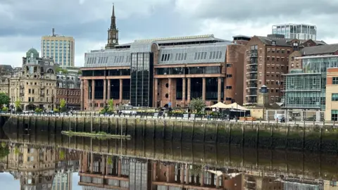 Newcastle Crown Court reflected in the River Tyne running in front of it. It is an imposing building made from smooth red stone with massive black windows and tall columns along its frontage.
