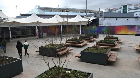 A paved outdoor area with about 10 visible banks of seating set around square flower beds, and a canopy over the entrance to a bus station in the background 