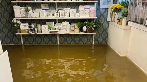 Sophie Ingram A shop that is completely flooded. Brown water fills the floor space and the shop shelves are full of small gifts such as picture frames and cards. The shop counter, which is painted cream, is on the right of the image and also has gifts - including a vase of sunflowers - sitting on it.
