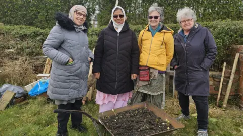 Aisha Iqbal/BBC A group of four women stand in an allotment. They are wrapped up warm. Two wear headscarves. There is a variety of gardening equipment around them, and they stand in front of a wheelbarrow loaded with soil.