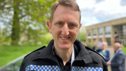 Chief Constable of West Mercia Police Richard Cooper , a man in a black police jacket is smiling at the camera. Behind him there are trees, a building and people who are blurred out