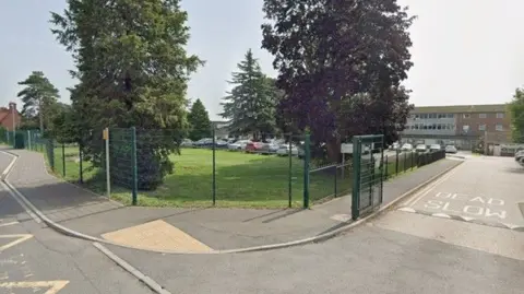 Google streetview image of Westfield Academy school. The school buildings are located behind a car park, with trees and grass in the foreground, and a green metal fence. 