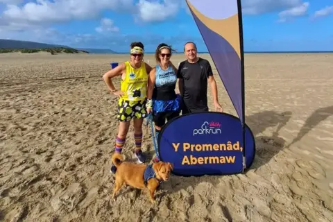 Jo Yarnall Two women and a man stand on a beach in running gear, with one of the women holding the lead to a small brown dog. They stand next to a purple Parkrun sign saying Y Promenâd, Abermaw