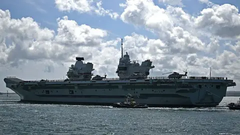 British aircraft carrier HMS Prince of Wales as it leaves Portsmouth Naval Base. It is during the day, the ship takes the entire picture. The waters are calm and the sky is blue but full of clouds.