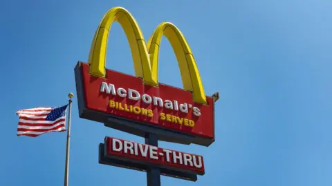 A McDonald's 'Golden Arches' sign against a blue sky with a US flag waving in the background to the bottom left