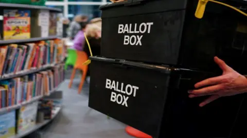 A person carries two ballot boxes stacked on top of one another through a library.