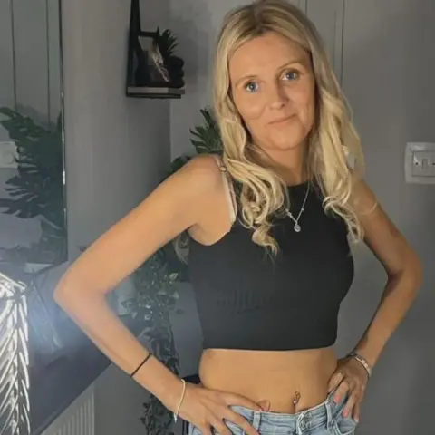 Carol O'Reilly Carol O'Reilly has long, blonde hair and is wearing a black crop top and faded blue jeans. She is standing by a mirror and is smiling at the camera