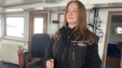 Ellie stands behind the wheel of the ferry, looking out to sea. She has brown hair and blue eyes and is wearing a black zip-up fleece.