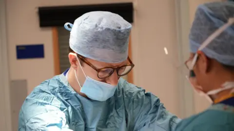 Shaun Whitmore/BBC Surgeon Nicholas Penney looking at one of the incisions he is making to accommodate the robotic arms. He is wearing a blue theatre gown and a blue face mask and is wearing his brown plastic rimmed glasses. He stands opposite an anethetist who is observing the patient.
