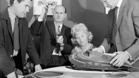 Getty Images A black and white photo of three men in suits and one woman examining a plastic model of a 'Stingray' submarine.