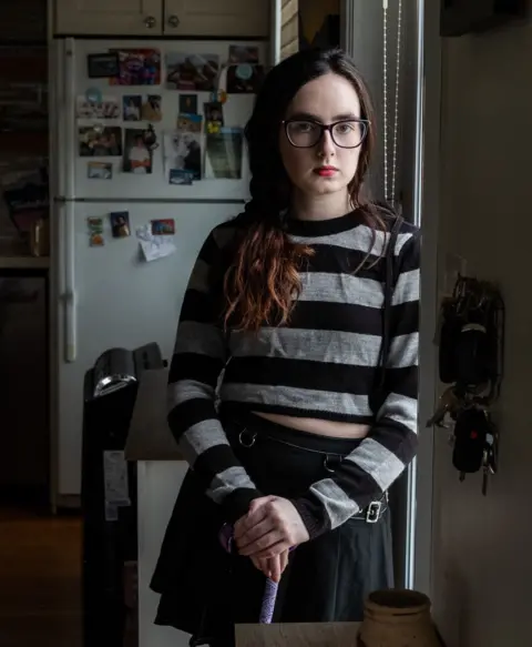 Gabrielle Cormier stands in her family living room, leaning on her cane, in the light coming in through the door. She is wearing dark rimmed glasses, a grey and black stripped long sleeved T-shirt and a black skirt.