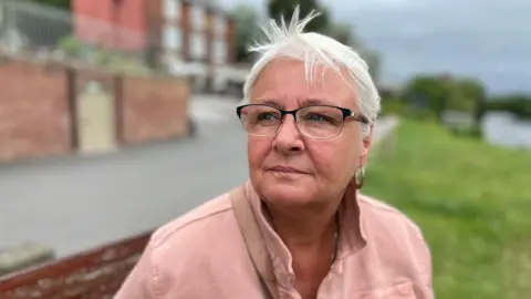 Helen is wearing a peach coloured jacked and glasses. She has short white hair and is sat on a bench near a canal.