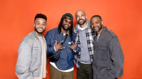 Four members of JLS posing for a photo with an orange background. They have their arms around each other. One member has both hands up in a "rock" hand pose.