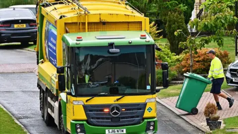 A yellow and green bin lorry driving towards the camera. A binman wearing a hi-viz jacket pushes a green bin on the right.