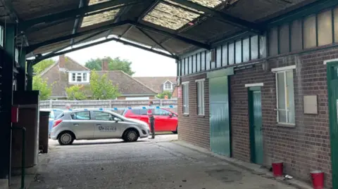 Health and Safety Executive A silver police vehicle is parked at the scene where the worker fell, with the area cordoned off. The image shows a roof over an empty area, which has an open end. The right side has doors and windows of a building.