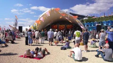 Festival attendees at First Light sit on the beach in front of a stage. Some are standing as they watch an act.